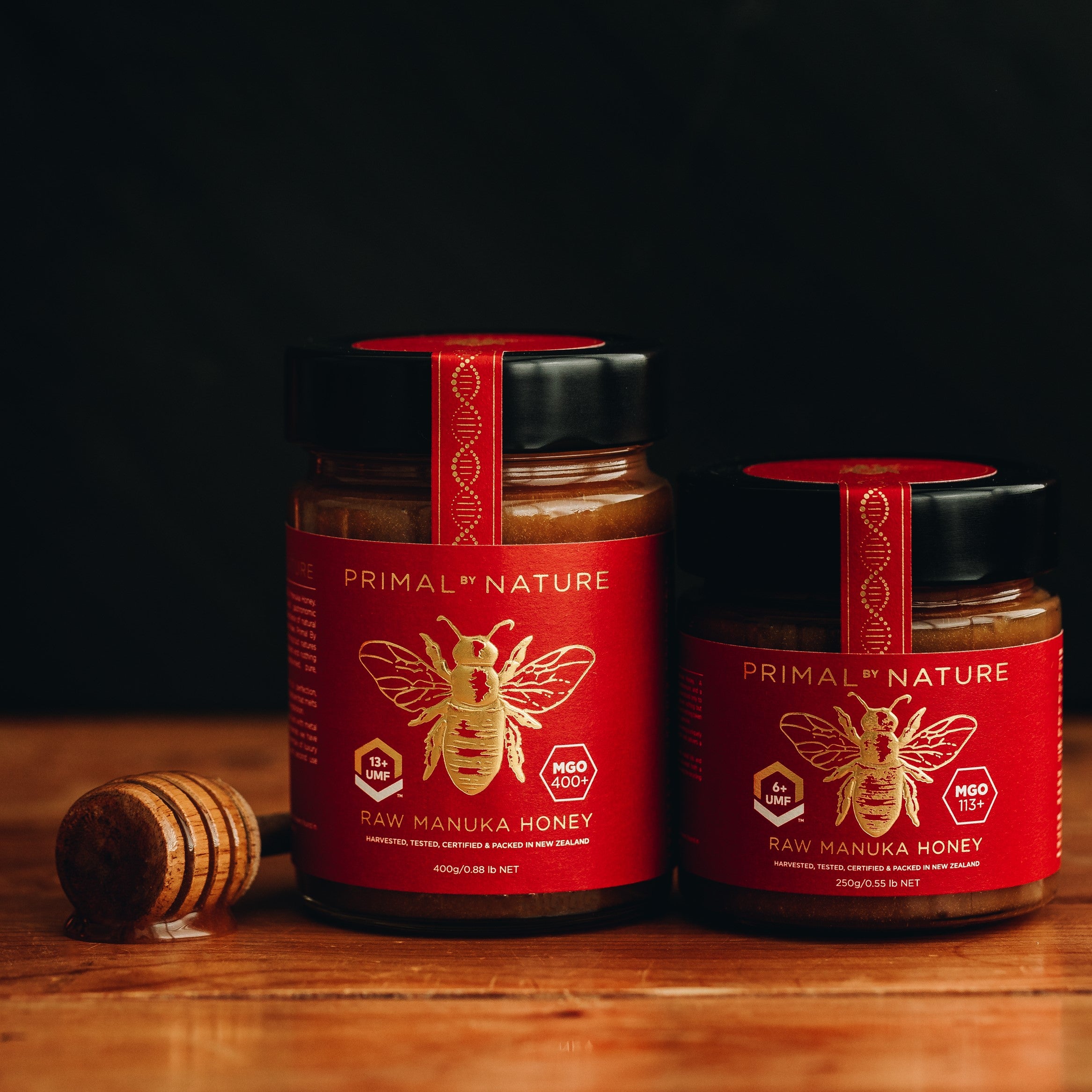 Primal By Nature Glass Jar Genuine 100% Pure Manuka Honey UMF 13+ MGO 400 400g next to 250g jar on a wooden cutting board with honey dipper