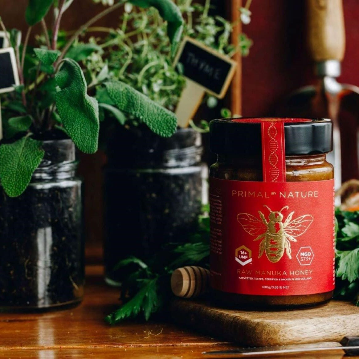 Primal by Nature Glass Jar Genuine 100% Pure Manuka Honey Jar UMF 16+ MGO 573 on a chopping board with reused herb pots from glass jars in the background